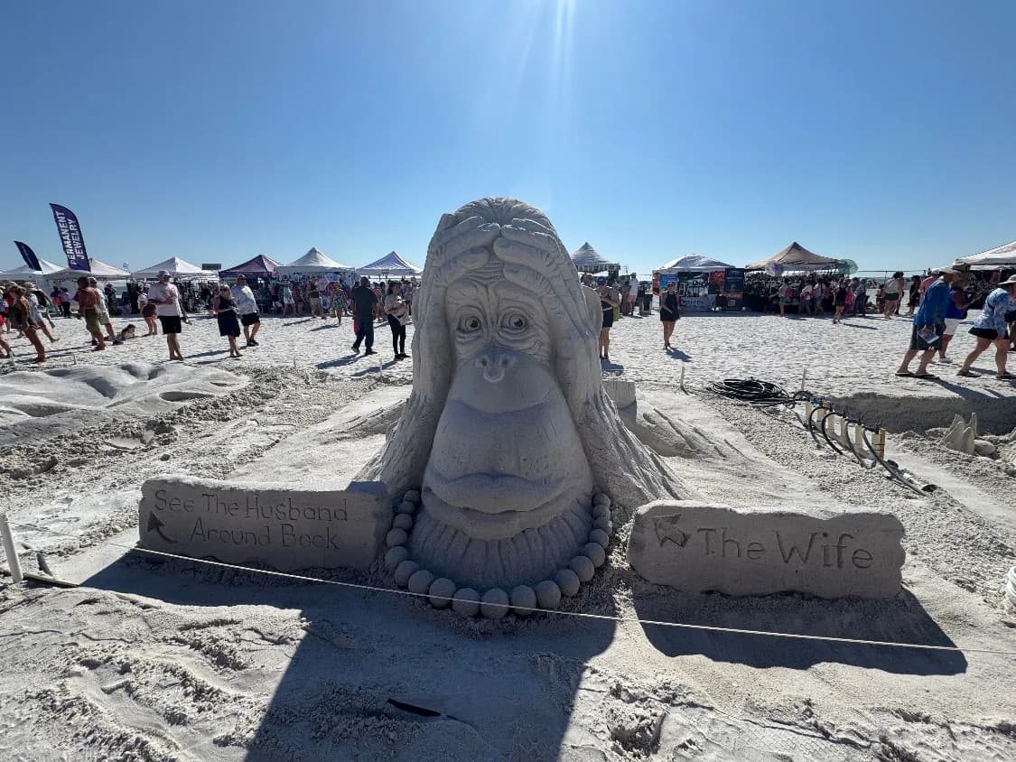 Sand sculpture of an orangutan wearing a bead necklace, with signs reading “The Wife” and “See the Husband Around Back,” at the American Sand Sculpting Championship on Fort Myers Beach