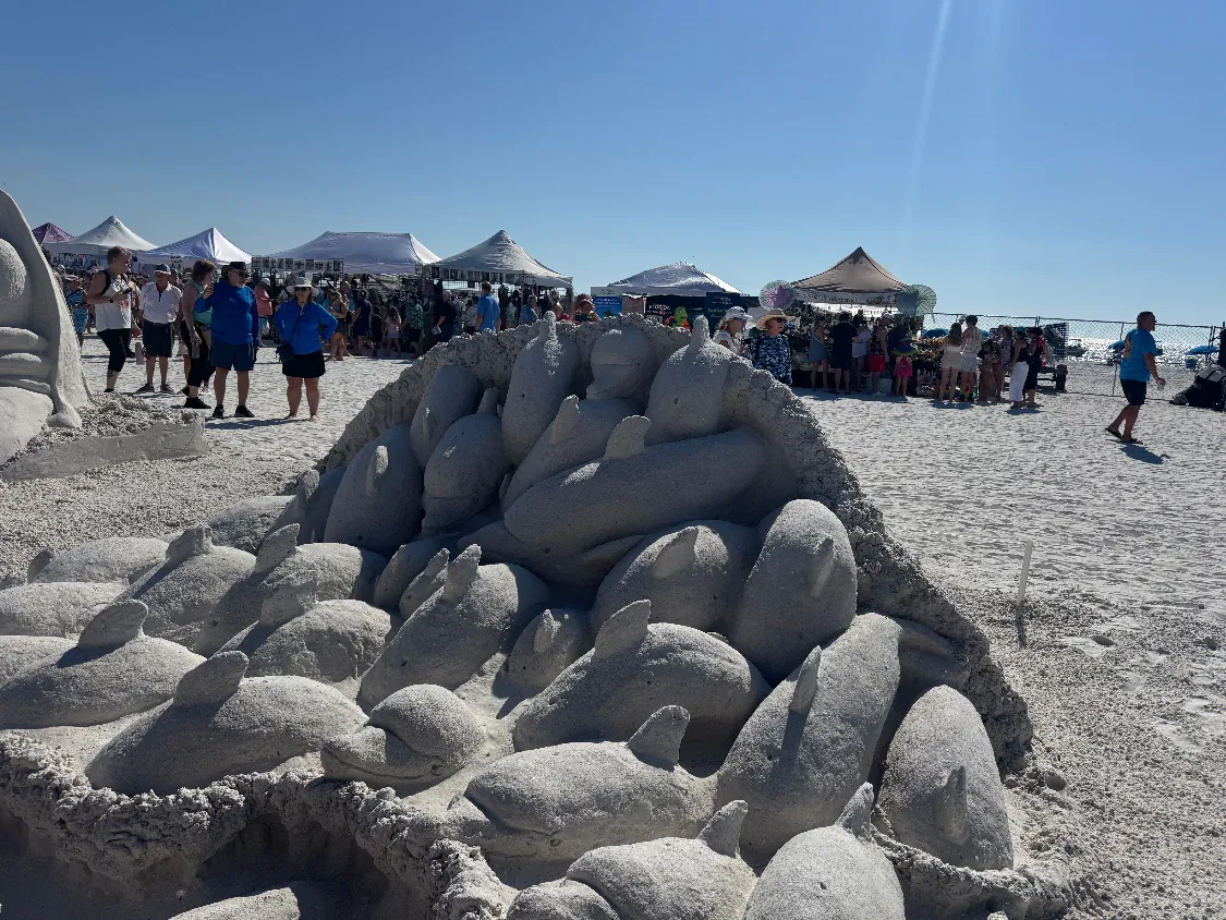 Sand sculpture of a pod of dolphins packed together inside a wave-shaped wall, with event tents and the Gulf of Mexico in the background