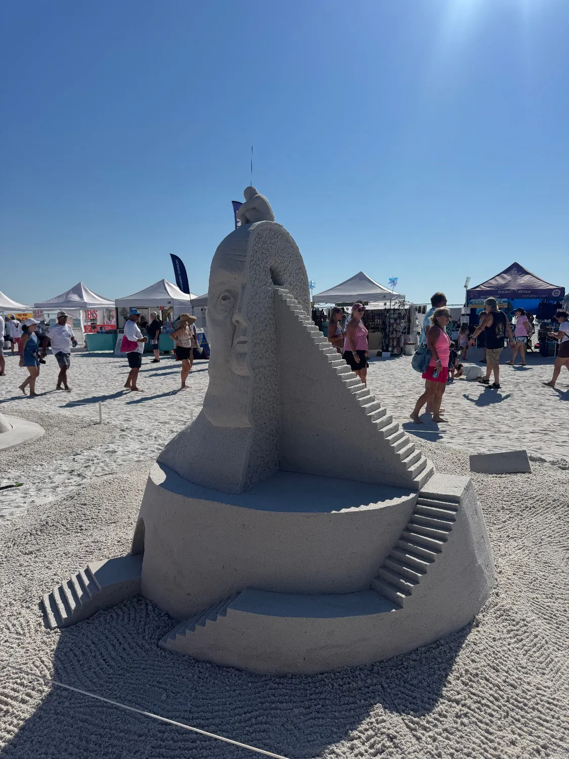 Sand sculpture of a large face with a staircase spiraling up its side to a small seated figure on top, at the American Sand Sculpting Championship