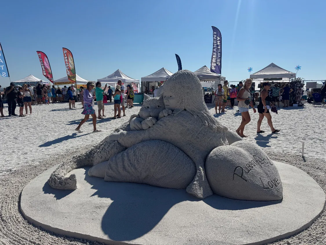 Sand sculpture of a girl hugging a cat beside a heart carved with the words “Purrfect Love,” with vendor tents and visitors on Fort Myers Beach behind it