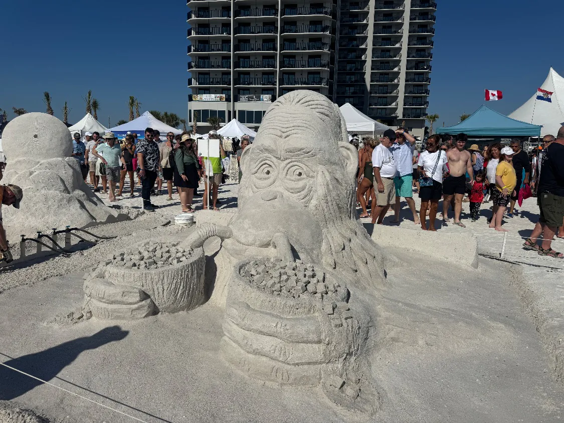 Sand sculpture of a monkey holding two baskets full of small sand cubes, with a crowd of visitors and the DiamondHead Beach Resort tower behind it